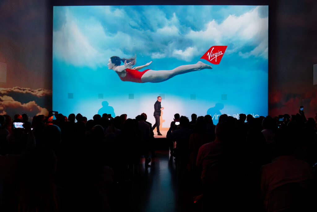 A giant screen with an image of a woman in a red bathing suit with her arms outstretched; she appears to be flying in the sky with Virgin's name and signature red on a tail near her feet. Virgin's CEO presents in front of the giant screen. Image: Virgin Atlantic