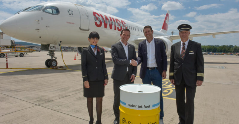 SWISS crew standing in front of an aircraft and a Synhelion barrel.