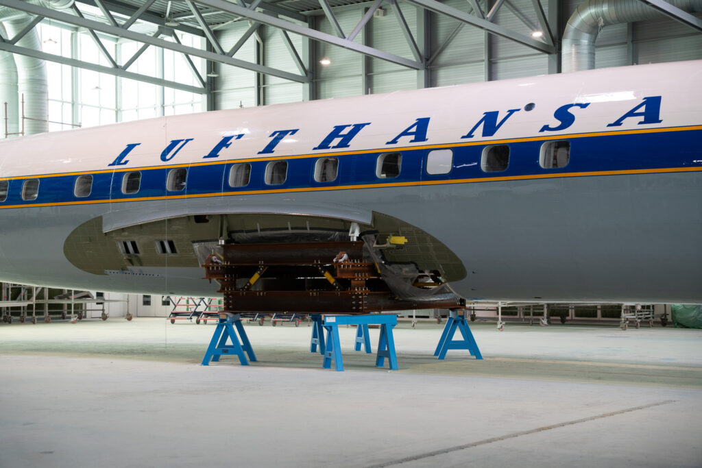 The word Lufthansa, - in blue lettering on a white backdrop - is painted on the side of the fuselage. The design looks very retroject style, with a thick blue stripe down the length of the aircraft, around the windows, surrounded by orange cheatlines. The bottom half of the aircraft is grey.