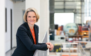 Nicole Dreyer-Langlet smiling towards the camera whilst an aircraft is being assembled behind her.