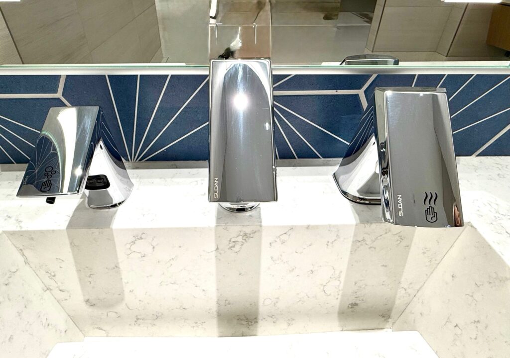 Restroom sinks have a blue tiling behind them and includes soap, faucet and dryer.