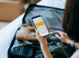 Over the shoulder view of young Asian woman using smartphone, checking digital flight ticket and boarding pass on device screen while packing a suitcase on a bed for a trip. Planning for travel. Travel and vacation concept
