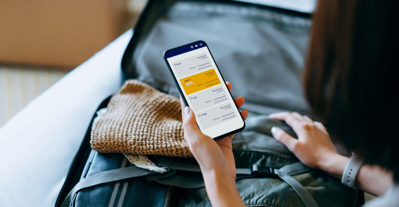 Over the shoulder view of young Asian woman using smartphone, checking digital flight ticket and boarding pass on device screen while packing a suitcase on a bed for a trip. Planning for travel. Travel and vacation concept