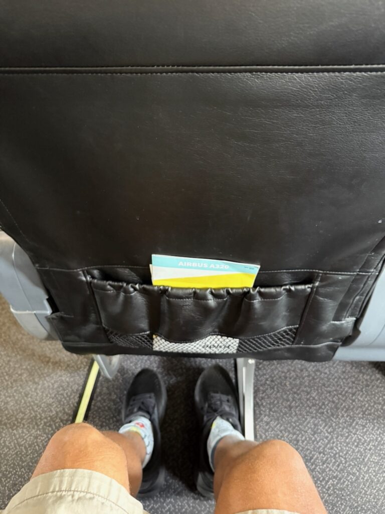A man's knees are seen a fair distance away from the aircraft seat in front of him.