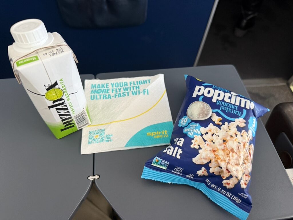 A BuzzBox Margarita and a bag of popcorn sit on a aircraft tray table.