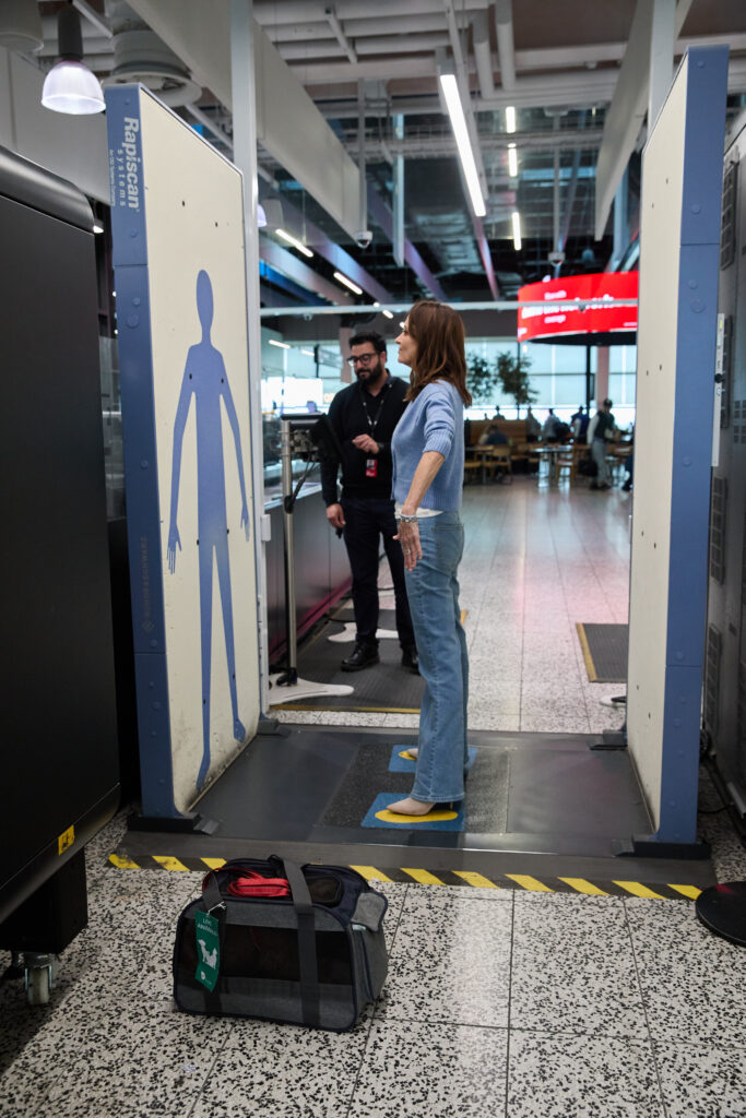 A passenger is being scanned at security while the pet carrier rests on the floor next to them.