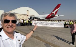 The Qantas A380 is at a hangar. Various members of the Qantas team are holding a banner and posing for a photo in celebration.