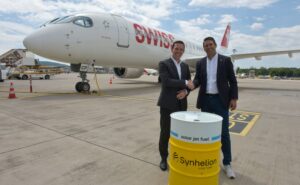 Jens Fehlinger, CEO of SWISS, and Philipp Furler, Co-CEO and Co-Founder of Synhelion, celebrate the long-term SAF offtake agreement while shaking hands behind a fuel barrel and in front of a SWISS aircraft.