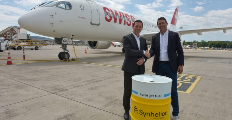 Jens Fehlinger, CEO of SWISS, and Philipp Furler, Co-CEO and Co-Founder of Synhelion, celebrate the long-term SAF offtake agreement while shaking hands behind a fuel barrel and in front of a SWISS aircraft.