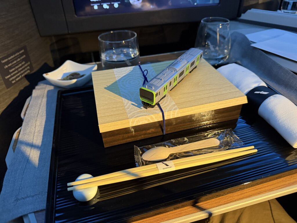 A boxed meal is displayed on the aircraft tray table including the branded wrapped chopsticks and a small train.