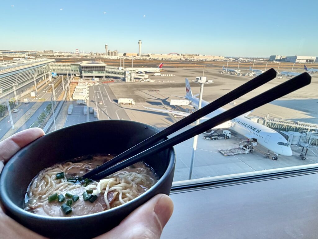 The author is holding up a bowl of noodles in front of a large window with a view of the ramp.