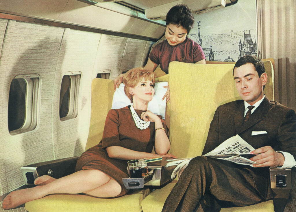 A vintage image of a Lufthansa cabin in what looks like the 1960s. A Lufthansa crew member positions a pillow beneath the head of a female passenger.
