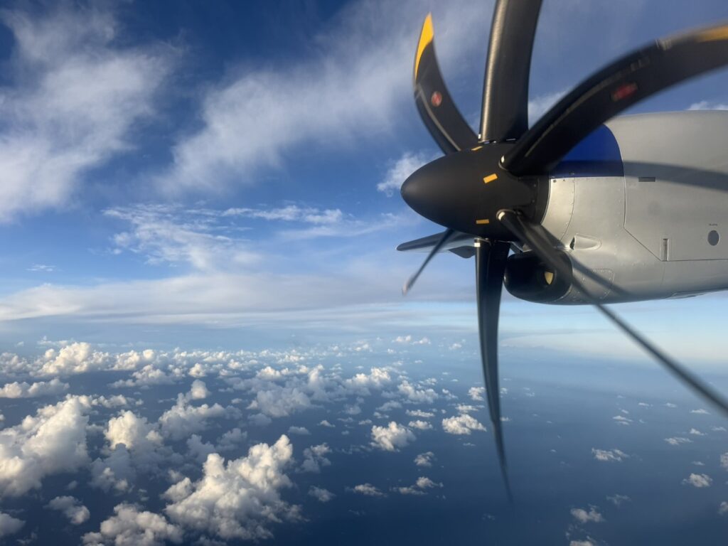 Looking out the window of the turboprop to see the turboprop engine up close and lots of fluffy clouds and a crisp blue sky above the clouds
