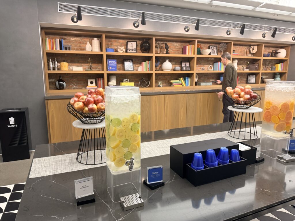 Two large basins of water with sliced citrus fruit sit next to a container of blue cups for self-service. 