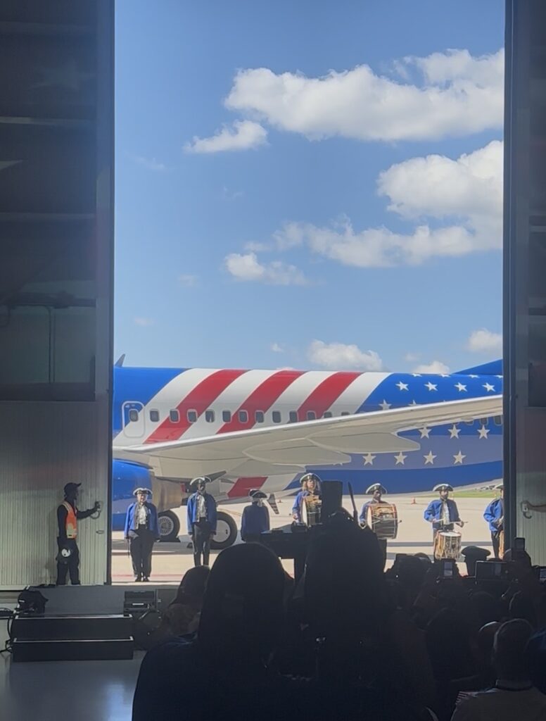 The widebody hangar door is opened to reveal the new livery, as a band plays American patriotic classics.