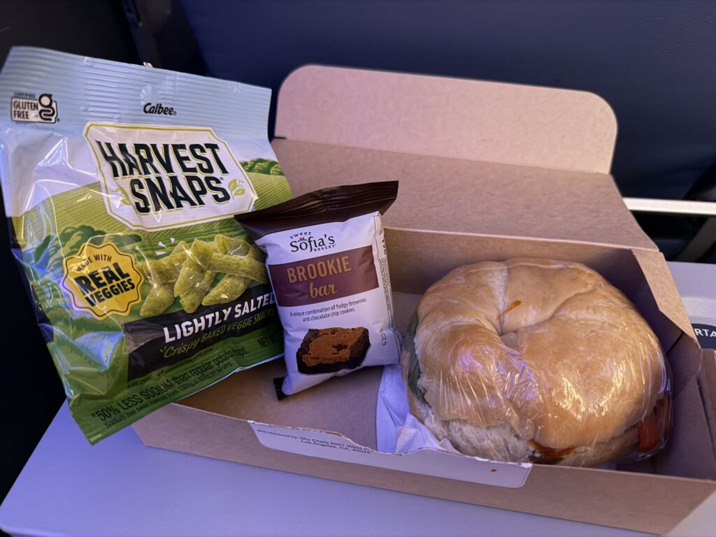 A Curry chicken croissant sandwich with a bag of baked veggie snacks and a brookie bar is served in Main Cabin on the American A321XRL, and laid out on the author's tray table