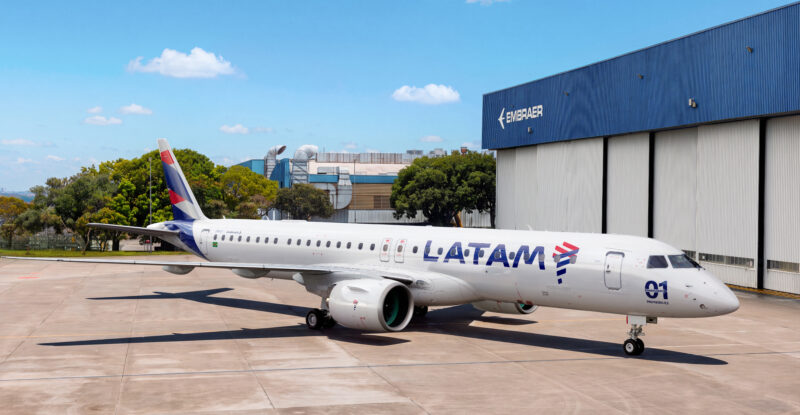 LATAM E195-E2 outside a hangar at the Embraer factory. The aircraft features a slimline antenna atop its fuselage to support inflight connectivity.