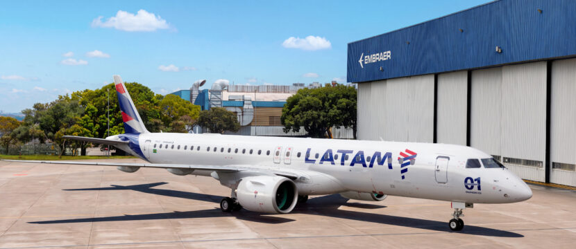 LATAM E195-E2 outside a hangar at the Embraer factory. The aircraft features a slimline antenna atop its fuselage to support inflight connectivity.