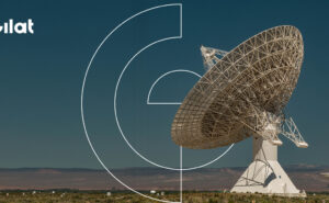 Gilat logo in the top left corner. The image shows a very large satellite dish in a large open space at night.