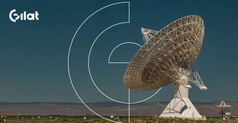 Gilat logo in the top left corner. The image shows a very large satellite dish in a large open space at night.