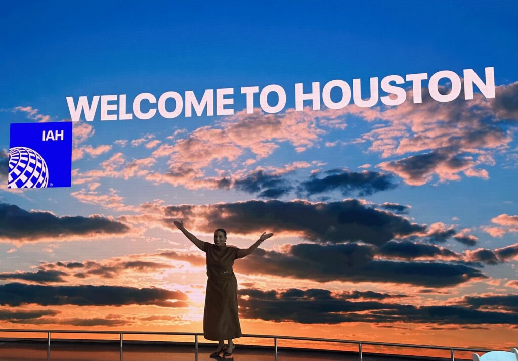 Large display says "welcome to Houston" with a woman standing in front of it, with her arms outstretched