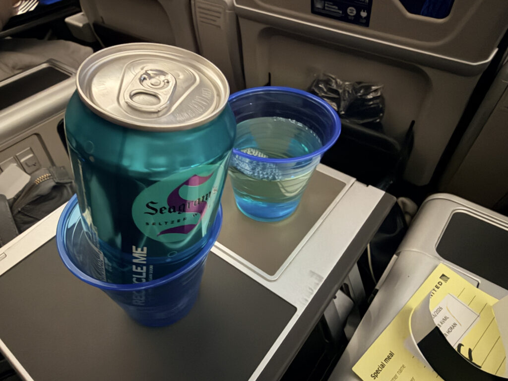 A can of soda is in a plastic cup on the tray table in Premium Plus aboard the United 787