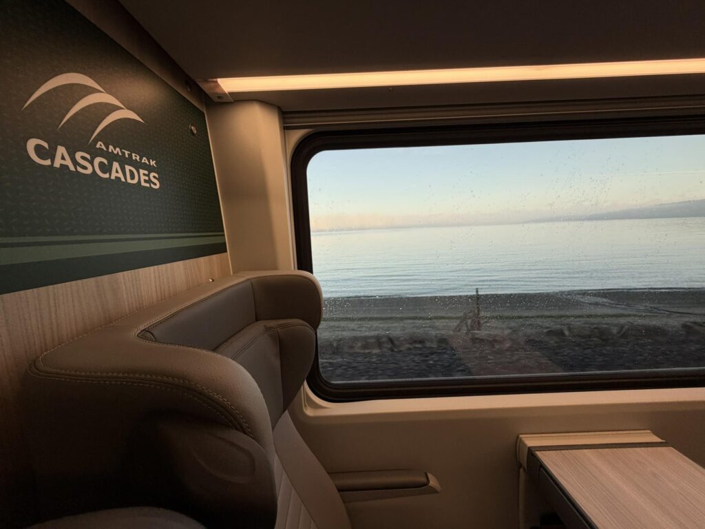 A comfy sseat, the words Amtrak Cascades against a green divider and a massive window showing a waterway on the Cascades route create a beautiful scene on board Airo.